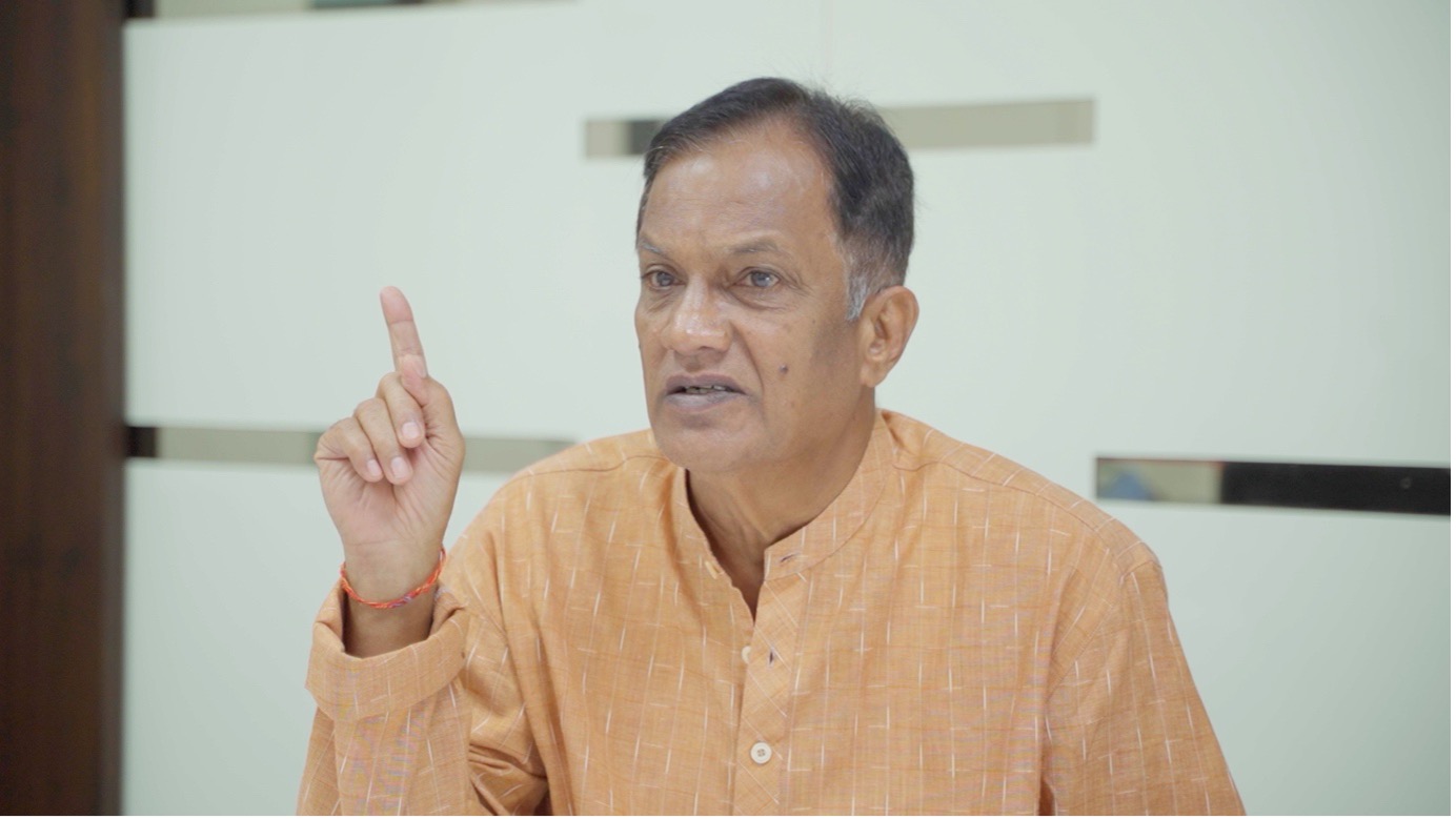 Jitendra Raul, resident and former village chief (sarpanch) of Navi Delwadi, speaking emphatically with one finger raised. He wears an orange kurta. A plain light-colored wall forms the background.