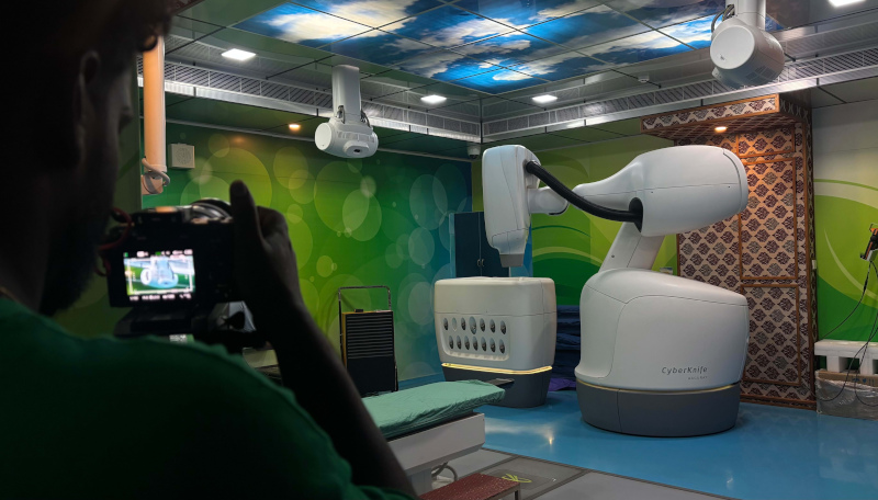 A cameraperson from the film crew records the CyberKnife radiation therapy system inside the treatment room. The machine and other clinical equipment sit beneath the blue ceiling displaying drifting clouds.