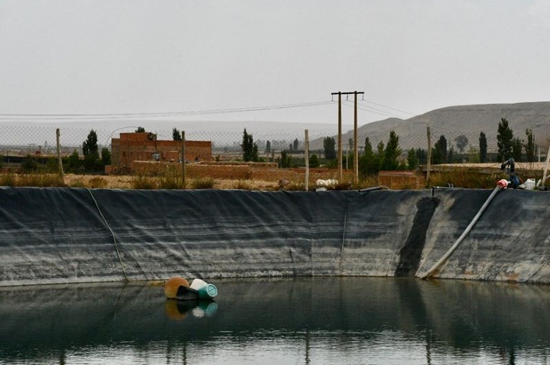An artificial embankment of black tarpaulin stretches across the image, against an arid gray landscape. The water level is low and white streaks are visible against the black tarpaulin.