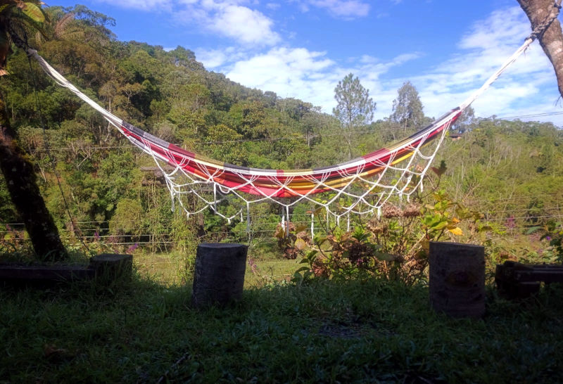 A colorful hammock, with red tones, is centered in the image and tied between two trees, in a forest setting, with a green hill and a blue sky with white clouds in the background.