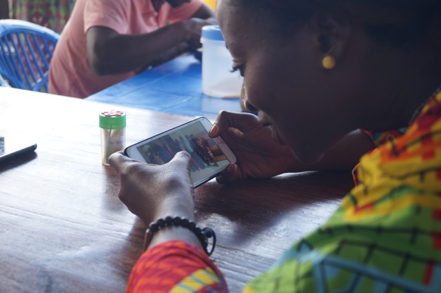 A black woman in a colorful top leans over her smart phone, looking at an image of a group of people.