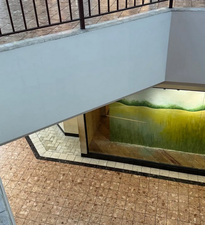 A color photograph of a shopping mall looking down taken from the second floor. Beige tiled floors and a landscape painting behind a glass display are visible. The bottom of a railing on the second floor is visible in the top of the image. No people are visible.