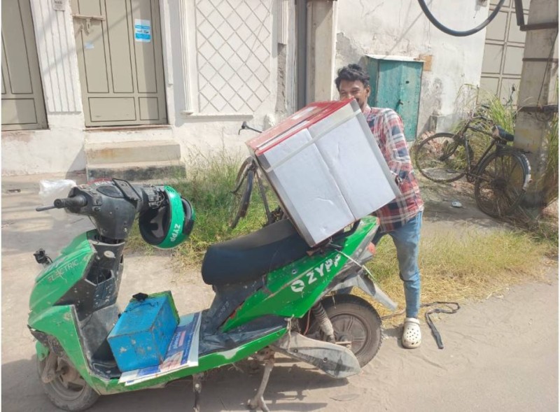 A man in a checked shirt and blue jeans is balancing a heavy white package on the back of his green electric scooter.
