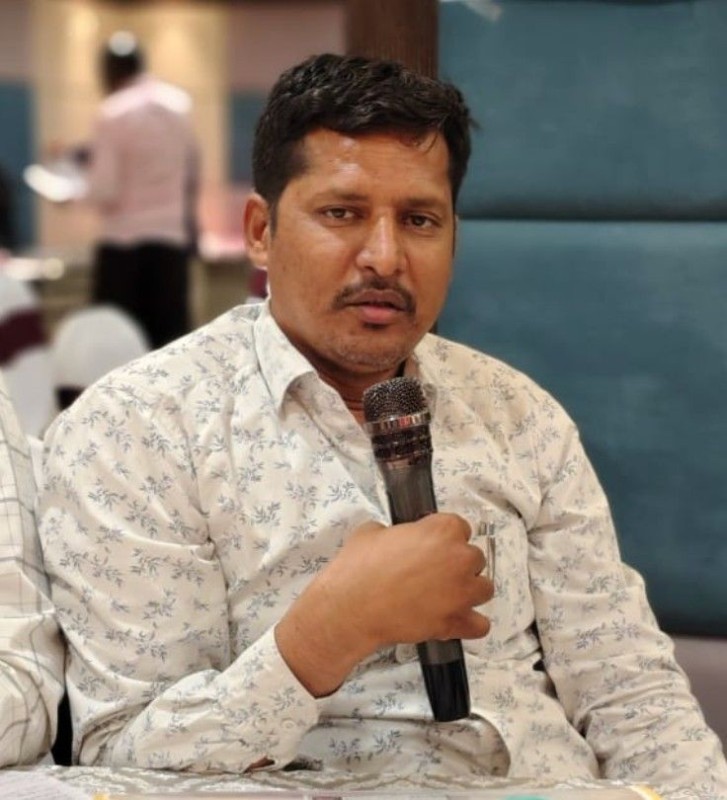A man with black hair and a white patterned shirt is holding a mike and speaking.