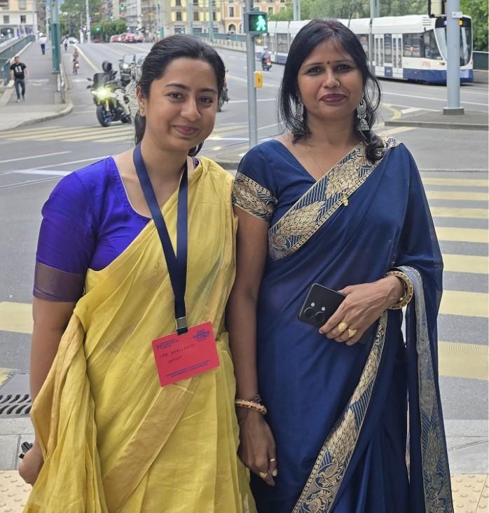 Two women, one in a yellow saree and the other in a blue saree, are standing next to each other and smiling at the camera. One of them has a conference lanyard around her neck. Image taken by author Isha Bhallamudi.