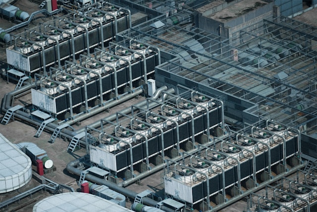 Aerial view of large rooftop industrial cooling units with rows of fans, interconnected pipes, and ventilation systems on the roof of a large industrial or commercial building.
