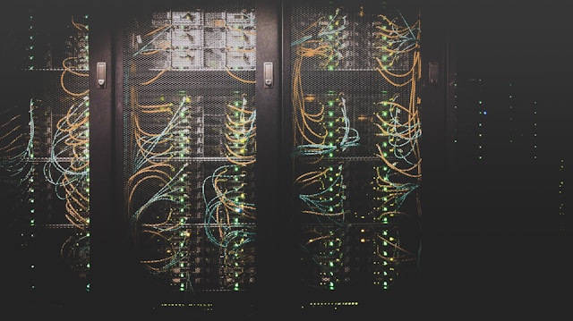 Several server racks filled with stacked servers and networking equipment, interconnected by dense bundles of cables with glowing indicator lights in a dimly lit data center.