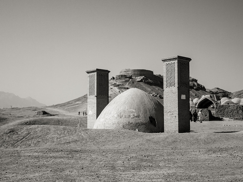 Black-and-white photograph of traditional domed buildings with two tall brick windcatcher towers, set in a dry, rocky landscape with a few people nearby.