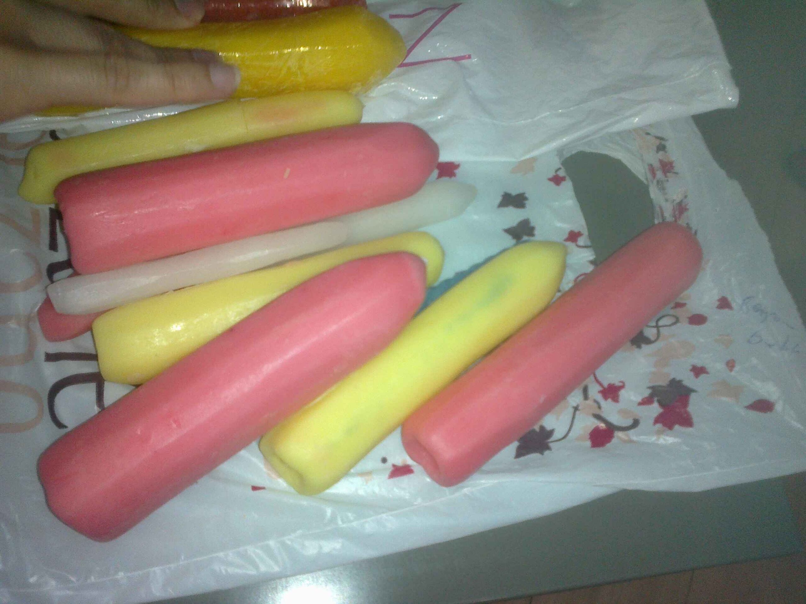 Yellow, pink, and white candles are laid out on a white patterned plastic bag on a table. A woman’s hand is sitting on a yellow candle on the left side of the image. The candles are all in different sizes and they all look carved and shaped. The white ones are the thinnest and the pink ones are the thickest. 