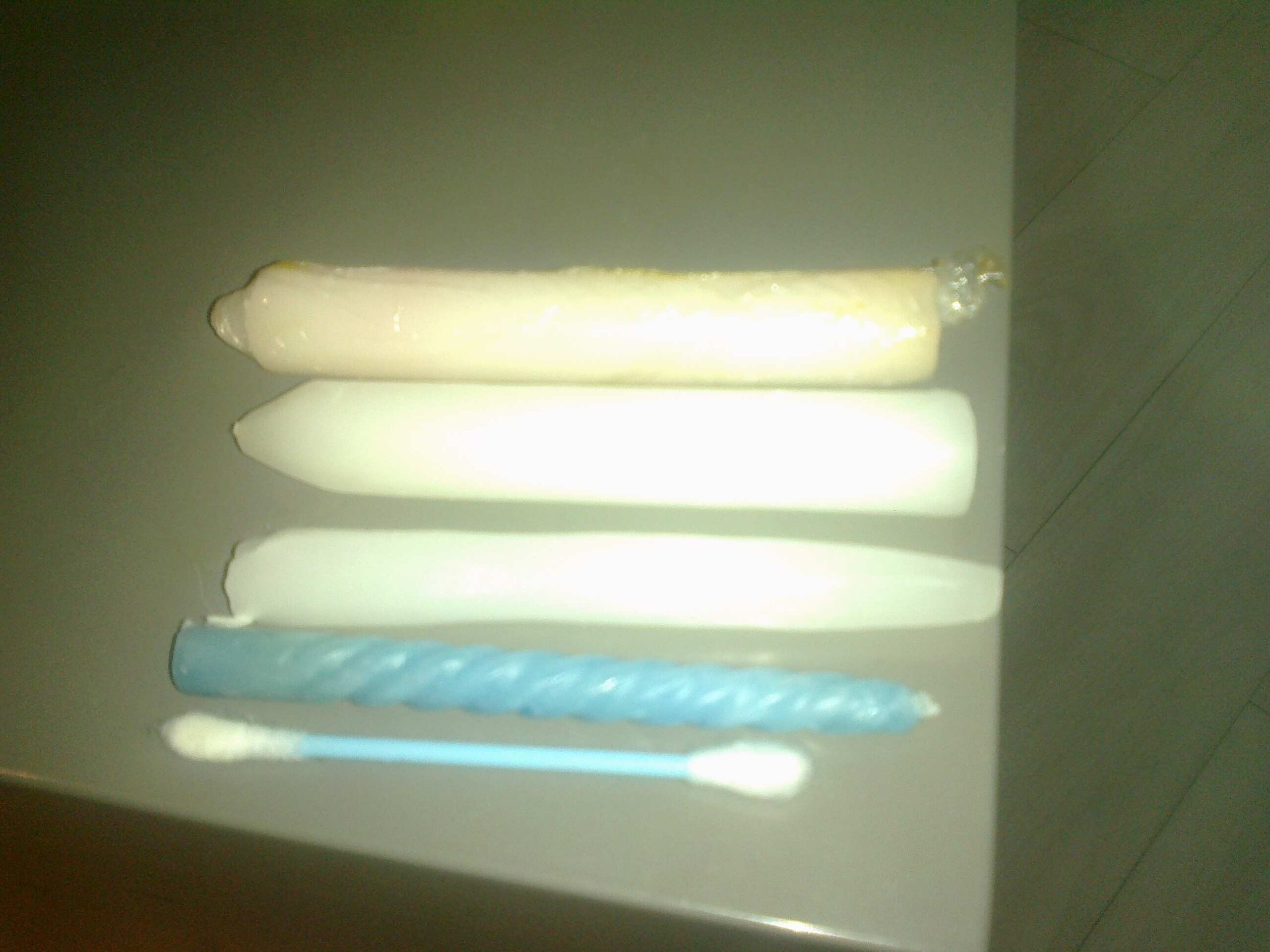 Pictured are three white candles, one blue birthday candle, and a cotton swab, laid out one below the other on a white table. All the white candles are melted into different sizes. Women generally start with the cotton swab and then move on to bigger candles as part of the therapeutic treatment for vaginismus.