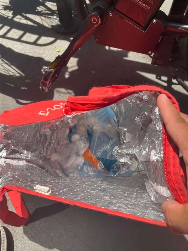 A coloured photograph of a hand holding a red bag with medicines in it, next to the shadow of a delivery bike