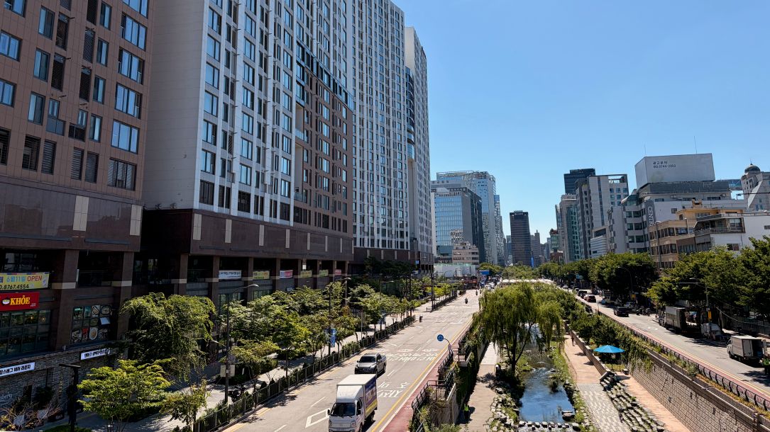 A highway runs through an urban area. In between the two directions of traffic, a stream flows. Large blocks of high-rise buildings tower on the left of the image.
