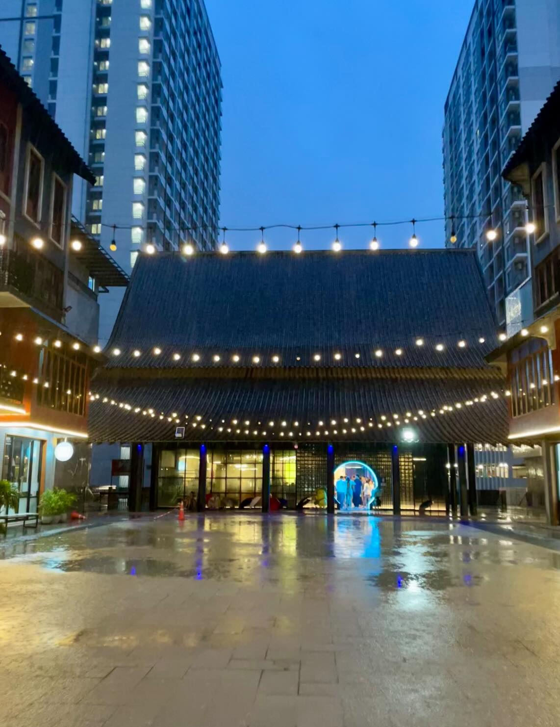 A building with a large dark tiled sloped roof, strung with warm string lights, stands at dusk in a plaza flanked by tall high-rise apartment towers. The wet pavement reflects the lights, and a small group of people gather near a glowing blue circular archway at the building's entrance.
