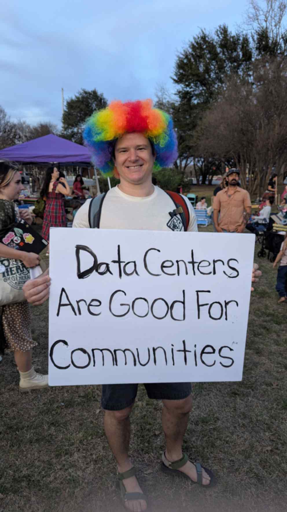 A person wearing a rainbow-colored wig holds a sign reading, “Data Centers Are Good For Communities,” at a community gathering.