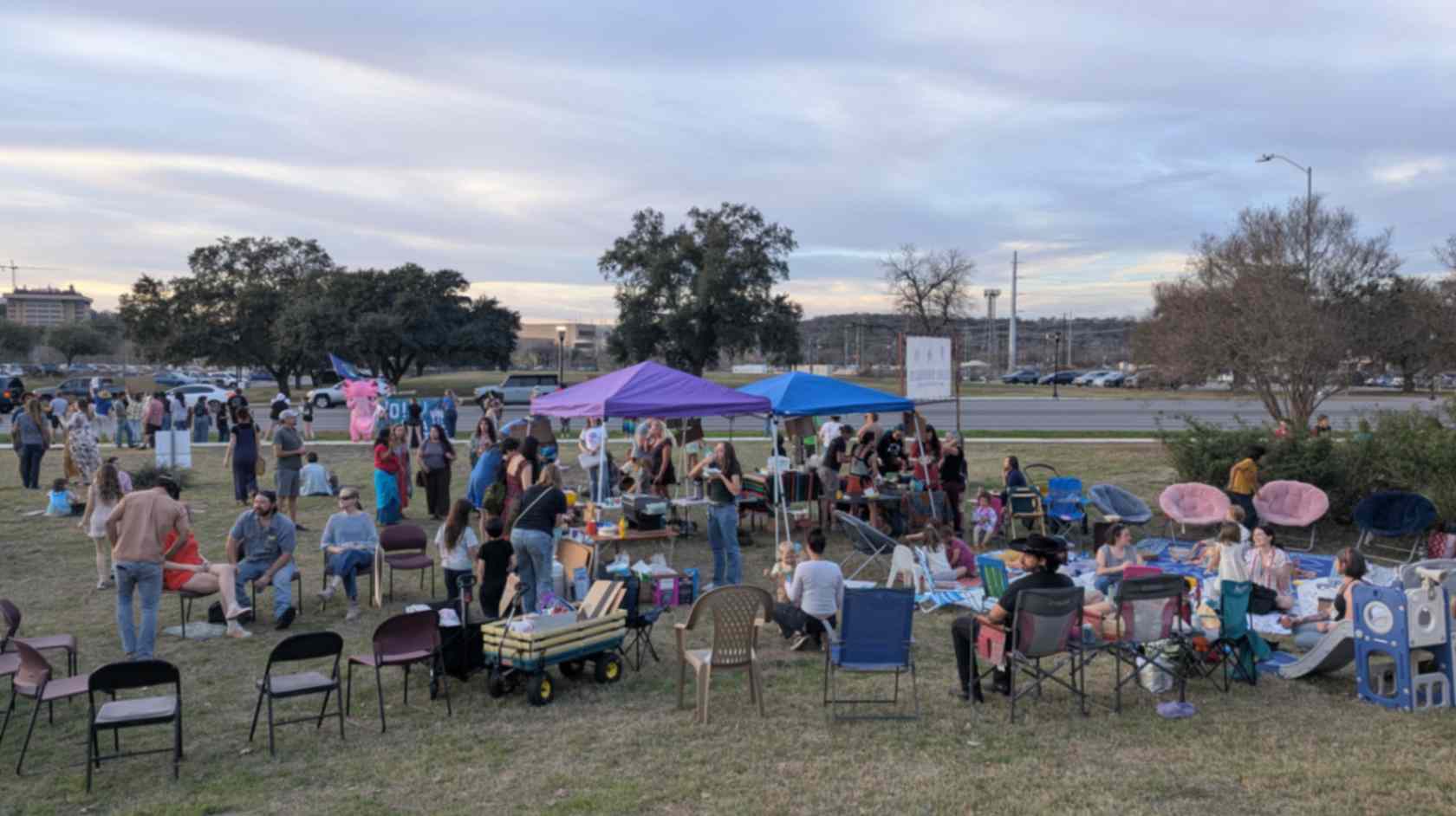 A large group of people gathers on a grassy field with tents, tables, chairs, and activities for children.