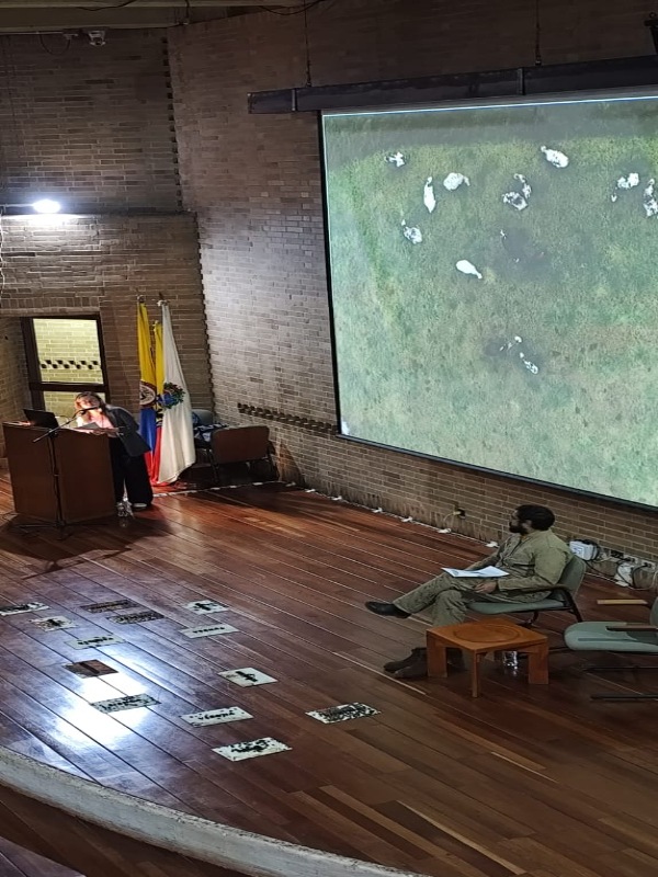 A man is seated on a chair, and a woman is speaking at a podium. Several images are displayed on the floor beside them, and a photograph of the green Colombian countryside is projected behind them.