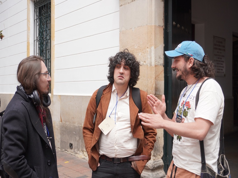 Three men—one wearing a black coat, another a gray jacket, and the third a T-shirt—talking on the street in front of a university.