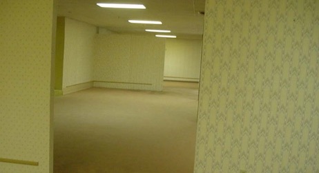 An empty, dimly lit carpeted hallway with patterned wallpaper and fluorescent ceiling lights.
