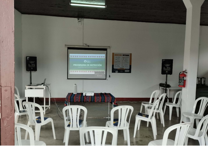 Meeting room arranged with plastic chairs in rows facing a screen displaying a presentation titled "Programa de Nutrición."