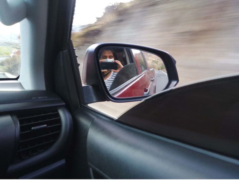 Masked person taking a photo in a car mirror. The scene shows part of the car interior and a blurred outdoor background. 