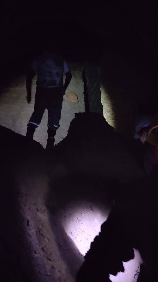 Two people inside a dark, earthen excavation, illuminated by flashlights. One stands beside a narrow trench, while another is partially visible in the foreground.