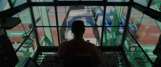 View from above the port with silhouette of port crane operator looking down on shipping containers on dock.