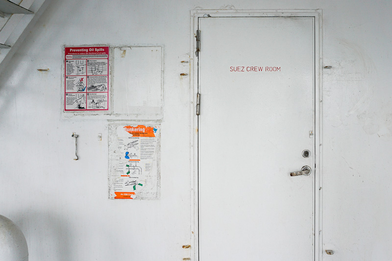 Photo of a closed door on the exterior breezeway of a cargo ship, painted white with red lettering stenciled onto it that reads, "Suez Crew Room."