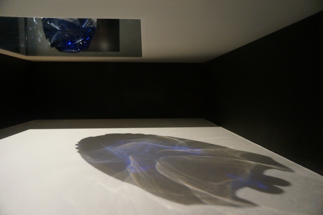 The colors and shape of the cut glass boat shaped indigo bowl are projected onto a large and expansive white tarp that lies on the floor.