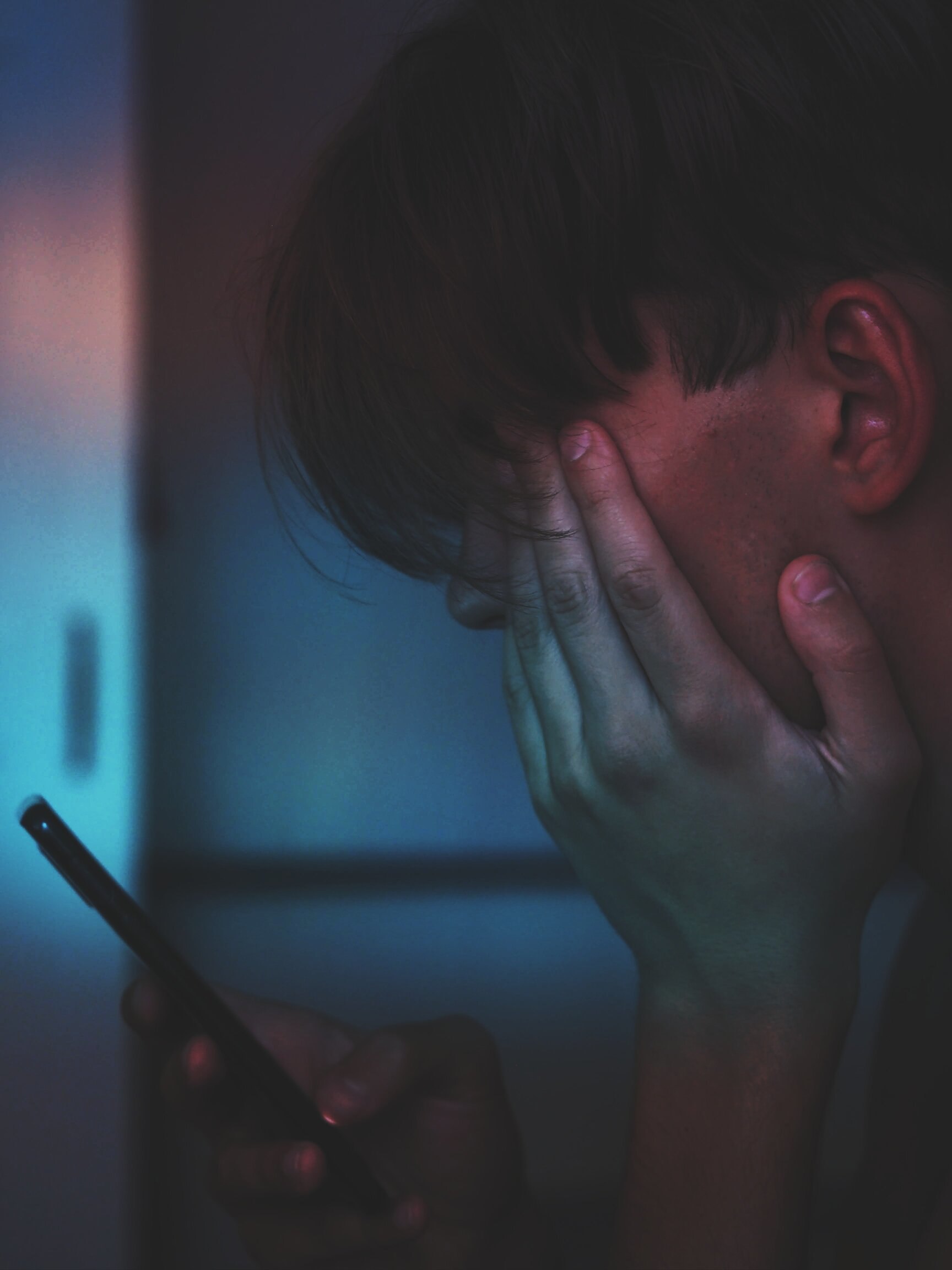 A young person with short hair that almost covers their eyes sits in a dark room looking at their phone, while holding their head