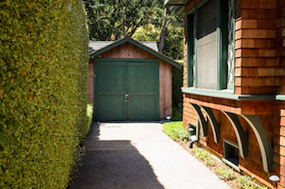 A driveway leads to a free standing garage with green doors locked by a padlock. On the left a thick row of hedges follows the driveway. On the right a bay window with green frame extends over a narrow strip of grass.