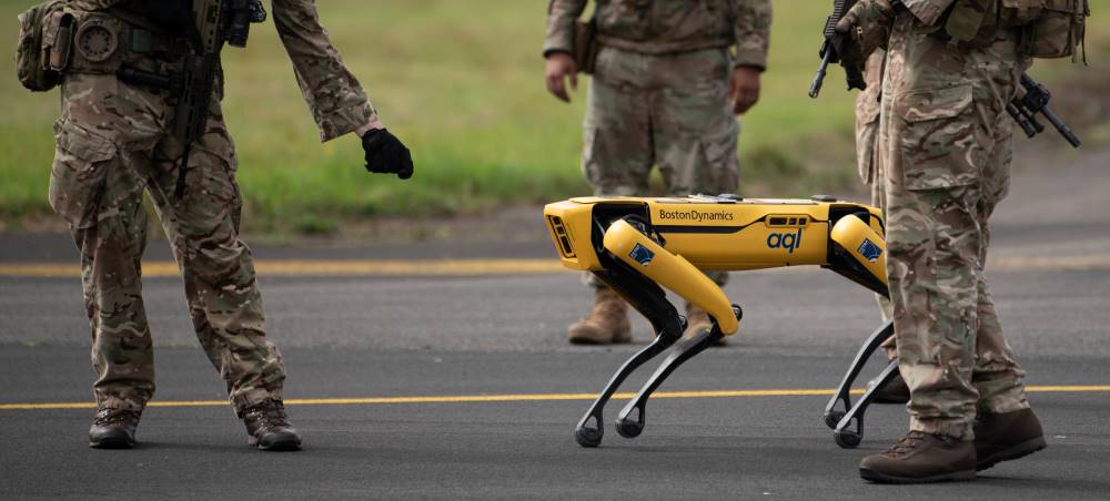 A yellow version of the Spot robot stands besides men in military garb