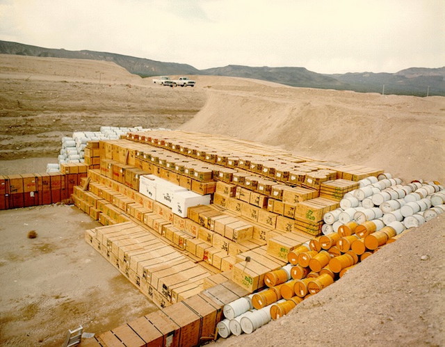 Large stacks of yellow, white, and red crates and drums are piled neatly in a massive sandy pit, set against a low mountainous landscape.