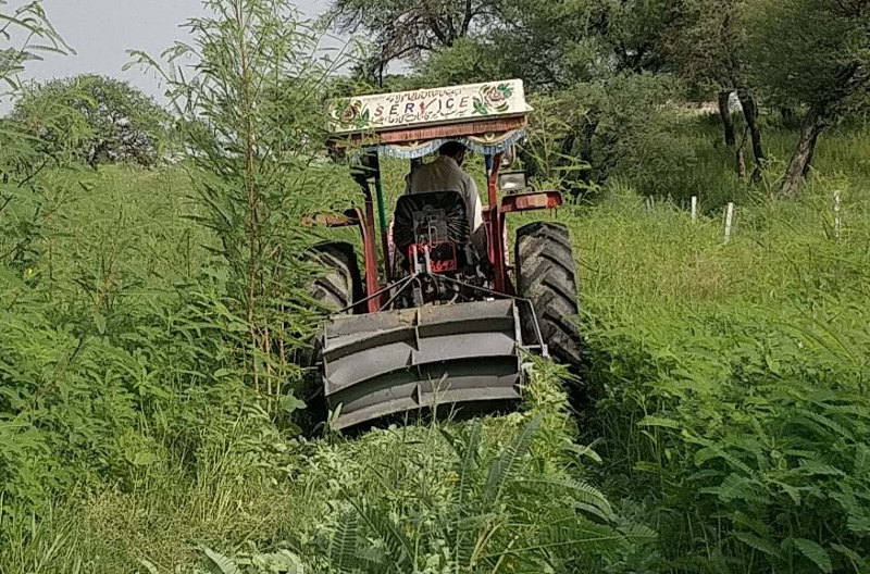 The image shows a green background, with a farm machine in the center. It has grey wheels. We can see the back of the driver in the photo.