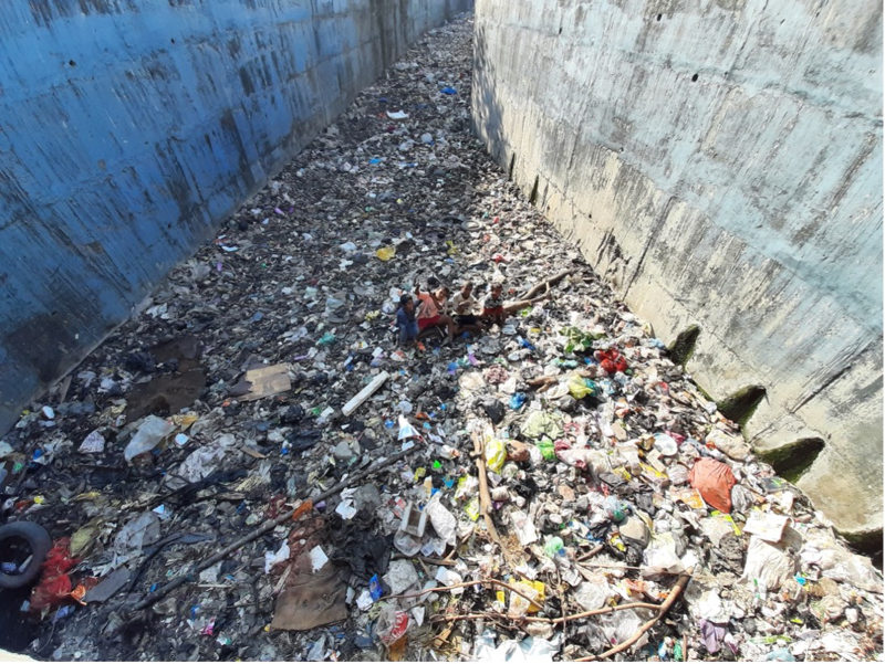 Image showing a vast expanse of scattered garbage and waste between tall concrete walls, with a few individuals sitting amidst the refuse.