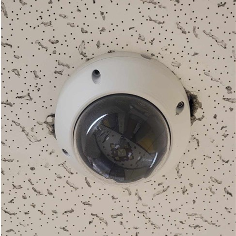 A dome surveillance camera mounted on a white ceiling tile. A hallway is reflected in the camera’s plastic housing.