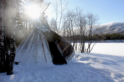 A conical dwelling made with sticks and a thick, snow-covered fabric is in the foreground of a snowy, wooded, and mountainous landscape. Sun shines through the trees.