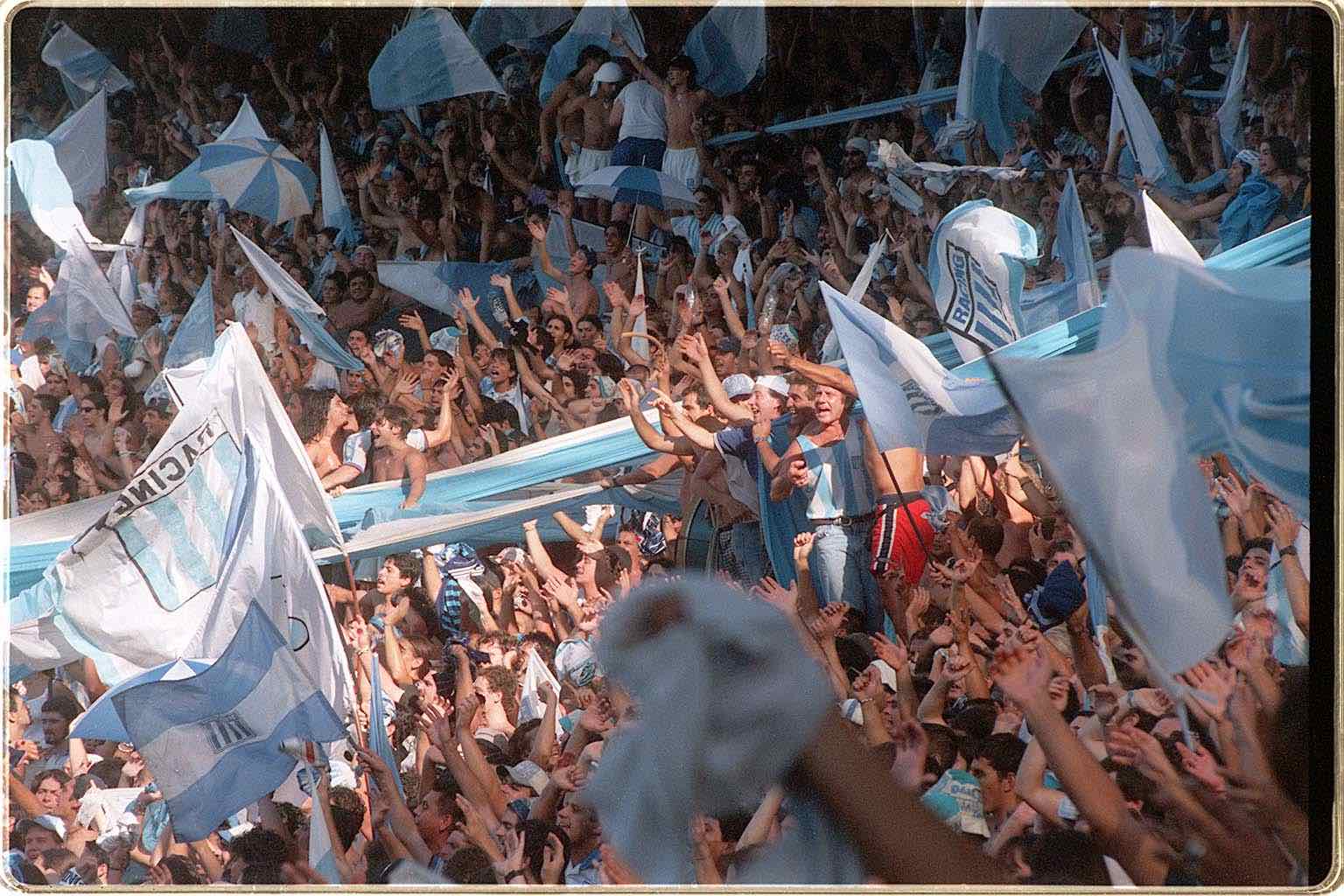 A crowd of people chant and have one or both arms raised. There are white and sky blue flags, some with the word Racing and Racing's logo, which is composed of sky blue and white vertical stripes.