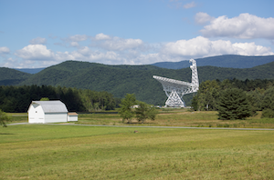 A white barn and a large white telescope sit in a grassy field with some trees around and tree-covered mountains in the background.