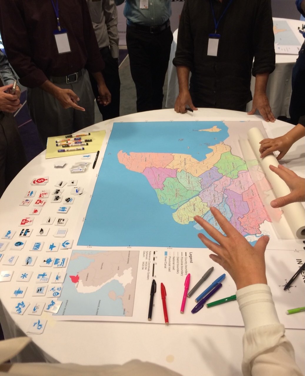 A map of the Mekong Delta oriented to the left is on a round white table. There are piles of small white squares with different icons organized into rows below the map. Colored markers and pens are also on the table next to and below the map. A person's hands are visible at the bottom left corner, while the bodies of others are also seen standing around the table.