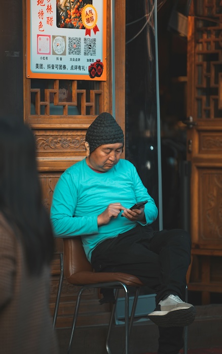 Person in turquoise sweater and knit hat sitting on a chair using a mobile phone, with traditional Chinese wooden architecture and signage in the background.