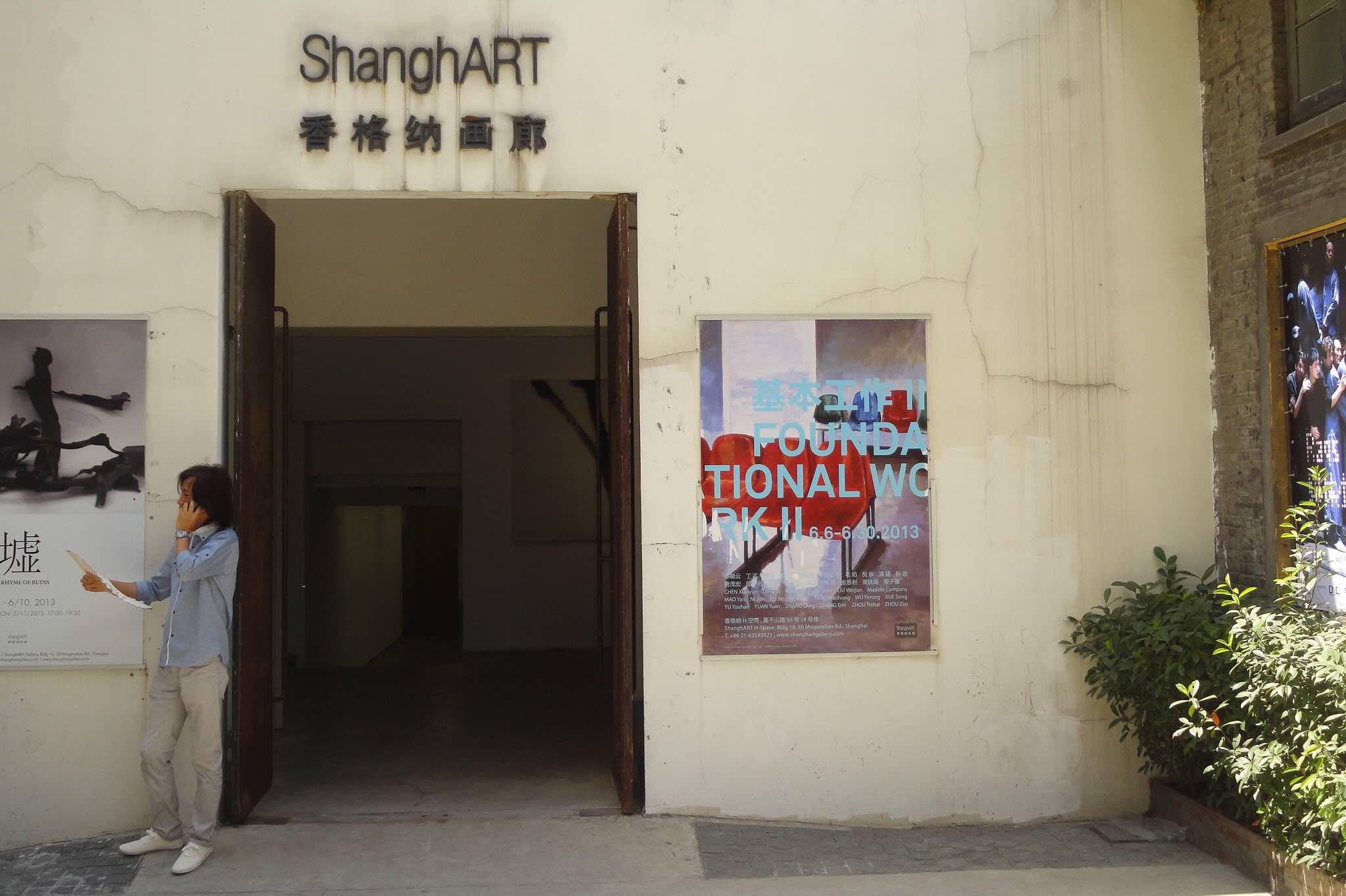 An individual stands outside of an off-white building that reads ShanghART with Canontese characters below it. The individual is holding a piece of paper in their right hand and a mobile phone to their ear with their left. On both sides of the large entrance to the building, posters for art exhibits are prominently featured.